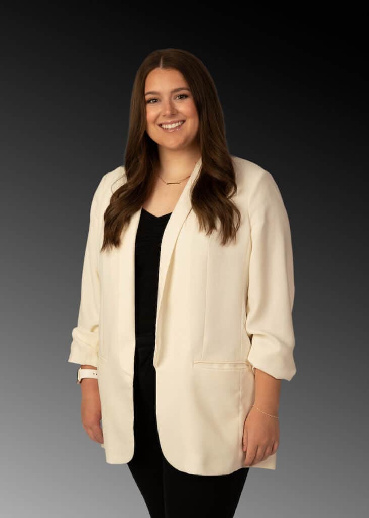 A woman with long brown hair poses for the camera in a black shirt and white blazer with her hands down at her sides while smiling.